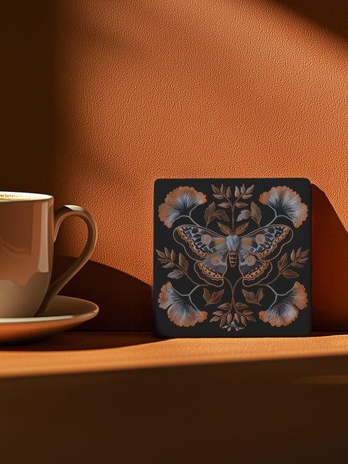 Square black ceramic coaster with detailed copper and blue moth illustration centered among symmetrical botanical elements including ginkgo-like leaves and delicate foliage.